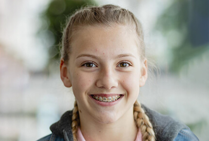 Smiling young girl with braided hair wearing dark jacket, indoor portrait with blurred background.