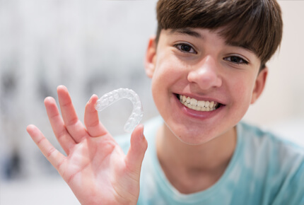 Smiling child holding clear orthodontic aligner in hand against blurred background.