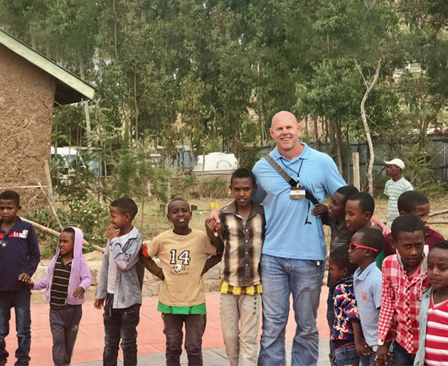 Man in blue shirt stands with group of African children outdoors near brick building.