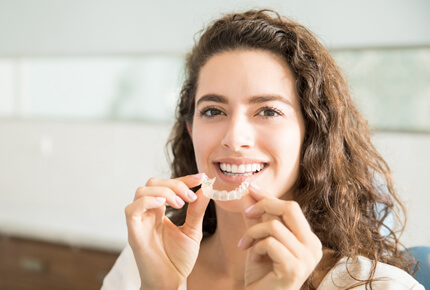 Woman smiling while holding clear aligner trays in dental office setting.