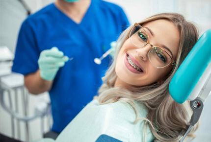 Young woman smiling in dental chair wearing glasses with orthodontist in blue scrubs behind her.