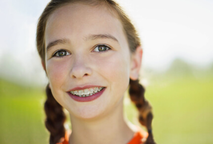 Young girl with braided hair smiling outdoors in a green field, displaying healthy teeth.