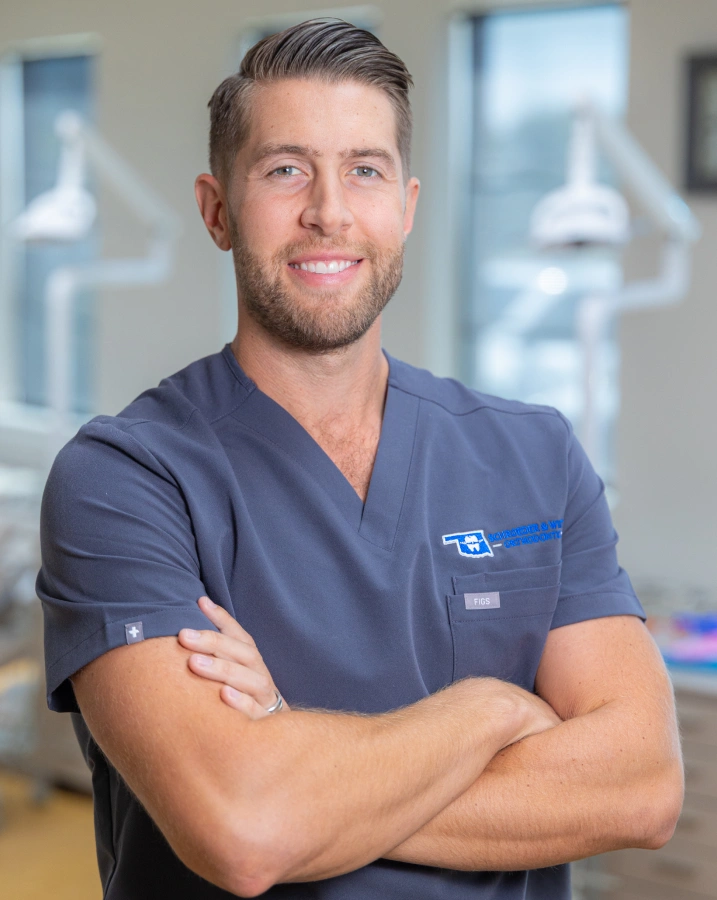 Male orthodontist in blue scrubs with arms crossed, smiling in modern dental office setting.