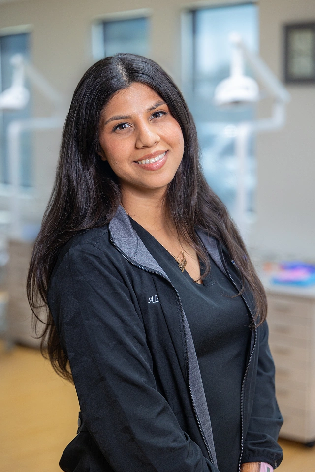 Young woman smiling at camera in modern orthodontic office setting wearing dark blazer.