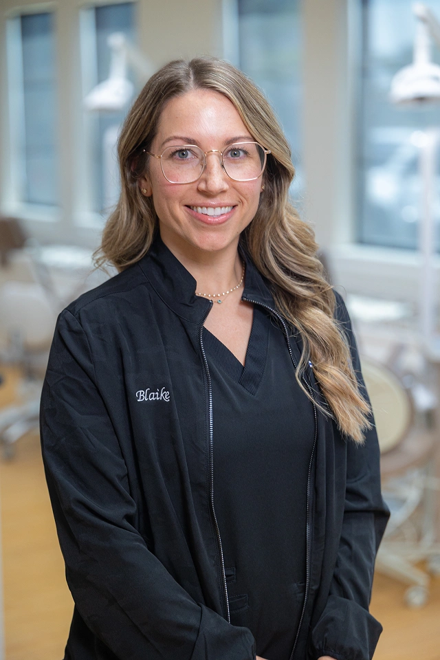 Professional woman in black jacket smiling at orthodontics office with windows behind her.