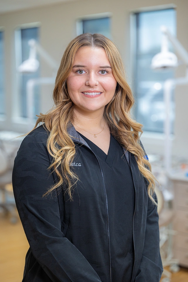 Professional portrait of a smiling woman in black blazer in modern office setting with windows.