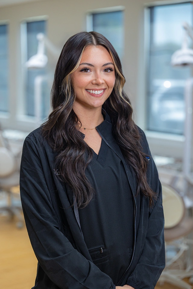Professional woman smiling at camera in modern office with windows, wearing dark blazer.
