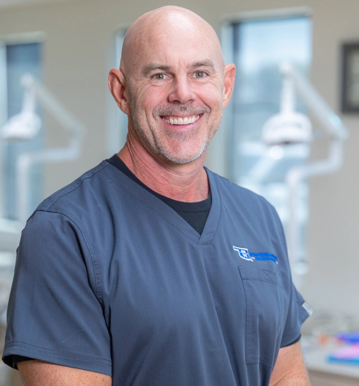 Male orthodontist smiling in clinical office wearing blue scrubs with company logo.